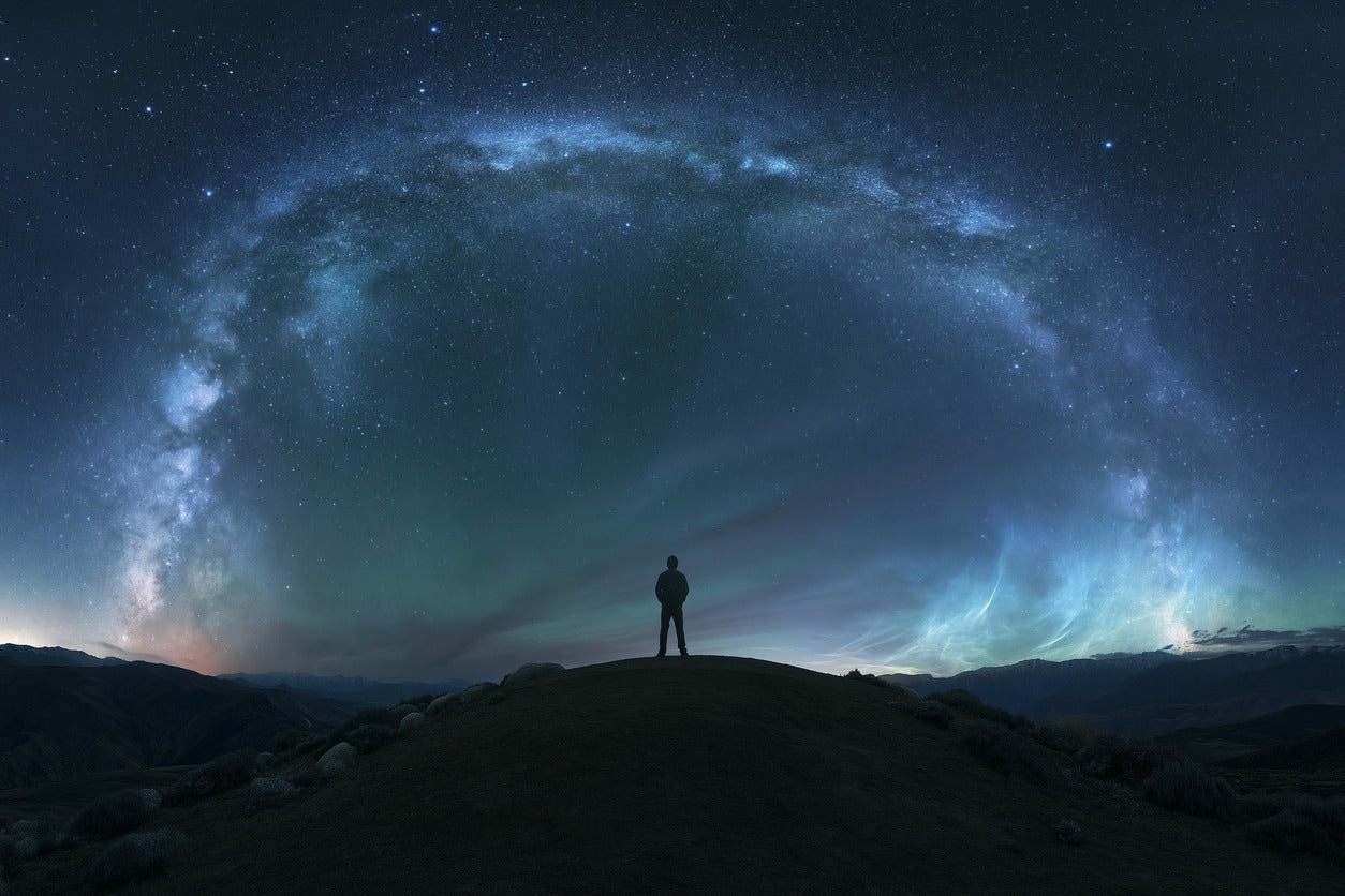 Lone figure on a dark hilltop beneath a sweeping arc of stars in deep blue night sky