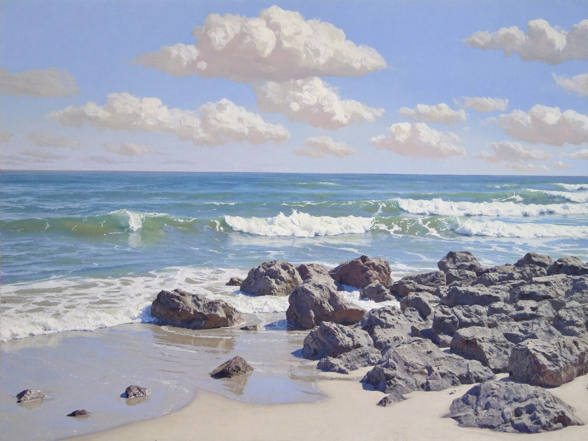 Rocky shoreline with rolling white surf, pale sand, and soft cloud banks over blue water