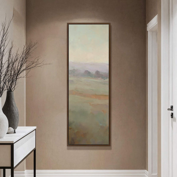 This vertical, unframed canvas print displays a soft, impressionistic landscape with a muted green and pastel palette, hung on a brown textured wall next to a console table with decorative.