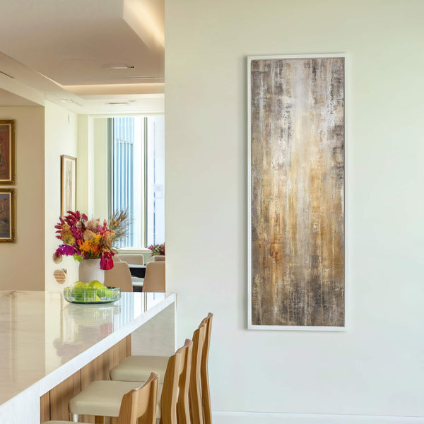 This vertical, abstract art print features a palette of earthy browns and grays with touches of gold, framed in white and displayed on a pale wall in a modern dining.