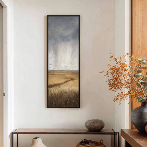 This vertical canvas wall art depicts a dramatic stormy sky over a golden field with tall grass in a realistic style, displayed on a white wall above a console table.