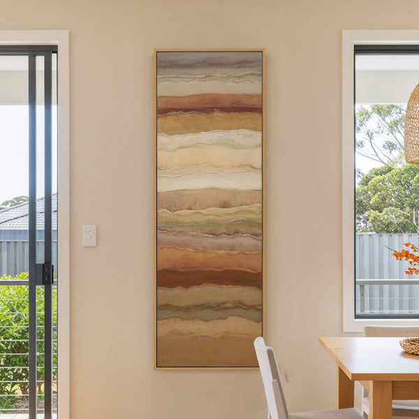 This vertical canvas wall art displays abstract horizontal bands in earthy tones of brown, beige, and gray, framed in natural wood, and hangs on a neutral wall in a room.