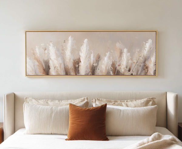 Framed pampas grass canvas wall art hung above a beige upholstered bed with layered neutral pillows in a cozy modern bedroom