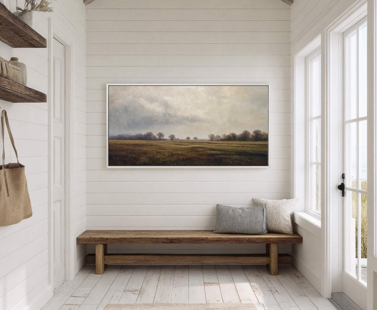 This horizontal canvas wall art features a muted landscape painting of a field with trees under a cloudy sky, presented in a white floater frame and hung above a wooden.