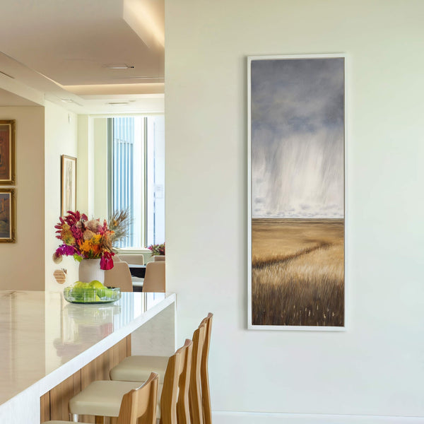 This vertical canvas wall art features a realistic landscape painting of a field under a dramatic, stormy sky, framed in white, displayed on a cream wall next to a modern.