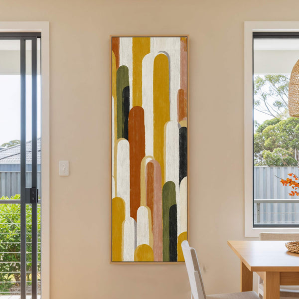 This vertical canvas print features an abstract pattern of rounded shapes in a warm earth-tone palette, set against a light beige wall, with a wooden dining table visible.
