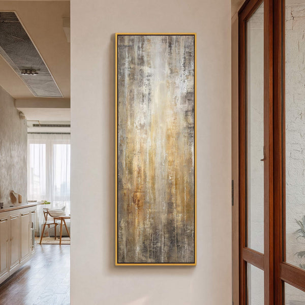 This vertical, abstract art print features a warm palette of beige, brown, and gold with textured vertical brushstrokes, framed by a thin gold floater frame, hanging on a beige wall.