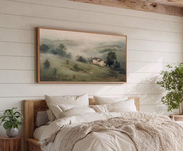 A horizontal canvas wall art print with a muted green and brown palette depicts a misty landscape with two houses and trees, framed in natural wood, displayed above a bed.