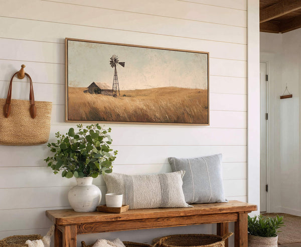 This horizontal art print depicts a rustic barn and windmill in a field of tall grass, rendered in a muted palette and impressionistic style, displayed on a white shiplap wall.