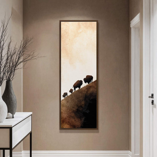 This vertical canvas wall art features a silhouette of bison walking up a hill in a warm, muted palette with a textured, abstract background. It is displayed on a beige.