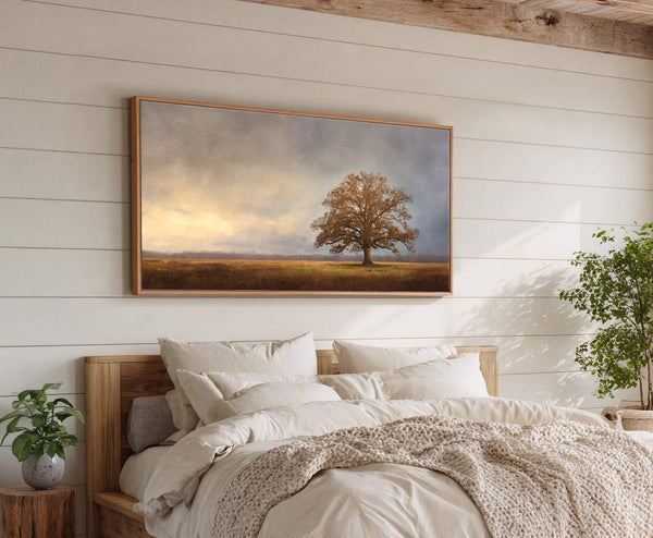 This horizontal canvas wall art features a lone tree in a field under a cloudy sky with warm light. The artwork is displayed above a bed with pillows on a.