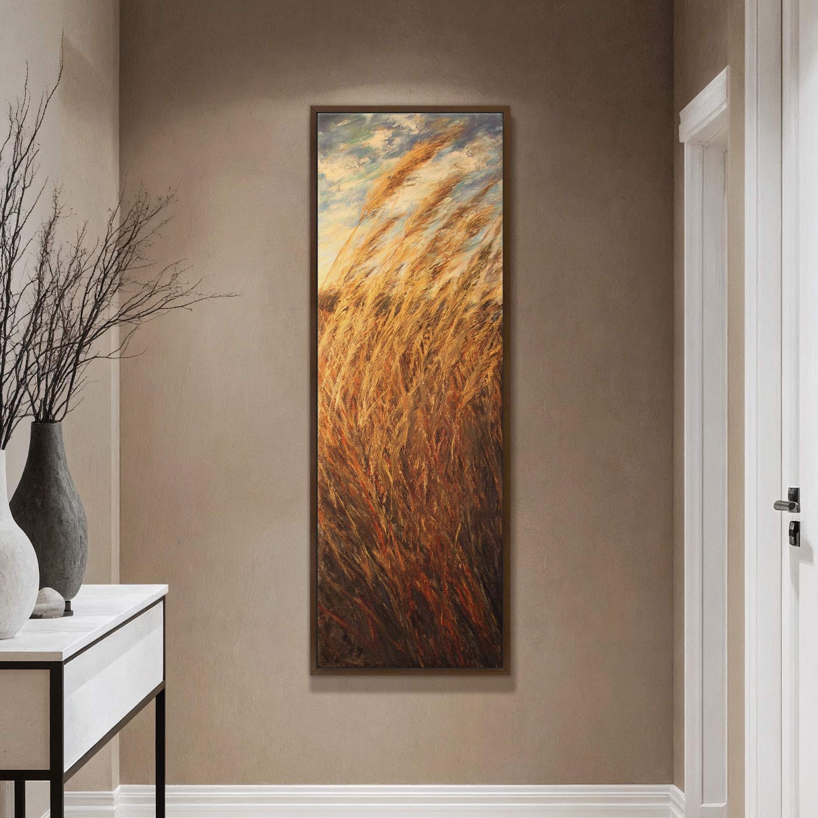 This vertical canvas wall art depicts tall, golden grasses in an impressionistic style with a palette of warm earth tones and sky blue. It hangs on a taupe wall above.