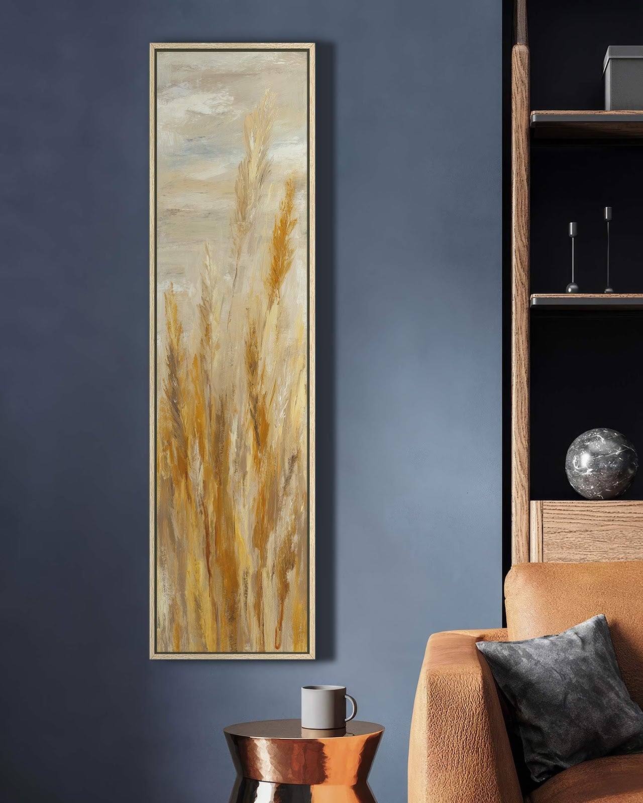 This vertical canvas art print features gold and beige impressionistic pampas grass in a natural floater frame, mounted on a blue living room wall beside a leather sofa.