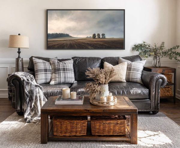 This horizontal canvas print features a muted palette depicting a cloudy sky over a tilled field and distant trees, framed in black, hanging above a dark leather sofa with plaid.