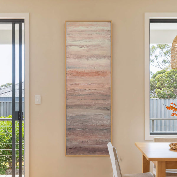This is a vertical canvas wall art with an abstract landscape palette featuring soft pinks, grays, and whites, framed in natural wood. It hangs on a neutral wall between two.