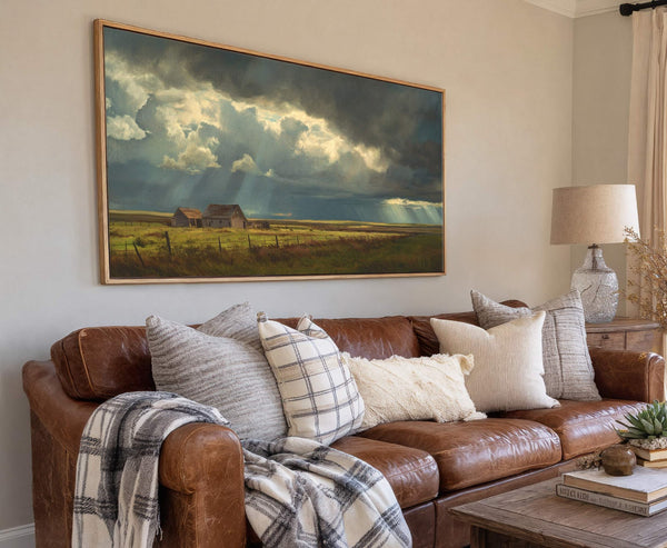 This horizontal canvas wall art depicts a dramatic stormy sky over a rural landscape with two barns. The palette is moody with dark blues and grays contrasted by bright sunbeams.