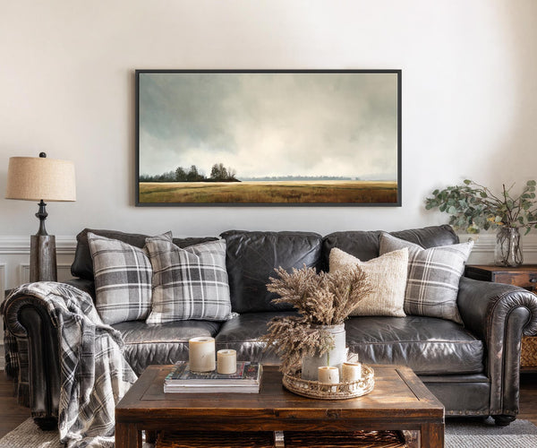 This horizontal canvas wall art depicts a muted, atmospheric landscape with a cloudy sky and distant trees over a golden field, framed in black. It hangs above a dark leather.