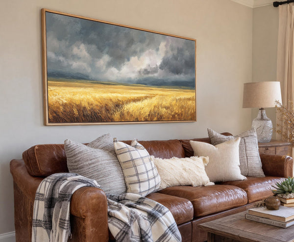 This horizontal canvas wall art depicts a dramatic landscape with stormy grey clouds over a golden field, rendered in an impressionistic style and framed in natural wood, hanging above a.