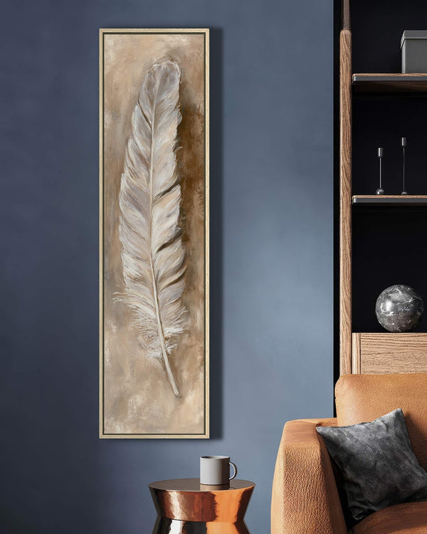 This vertical canvas wall art depicts a textured feather in neutral tones, displayed in an oak floater frame on a dark blue living room wall beside a tan sofa.