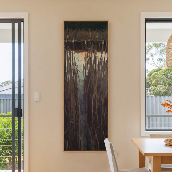 This vertical canvas wall art features a moody, impressionistic depiction of cattails by a body of water, framed in natural wood. The artwork is displayed on a light beige wall.