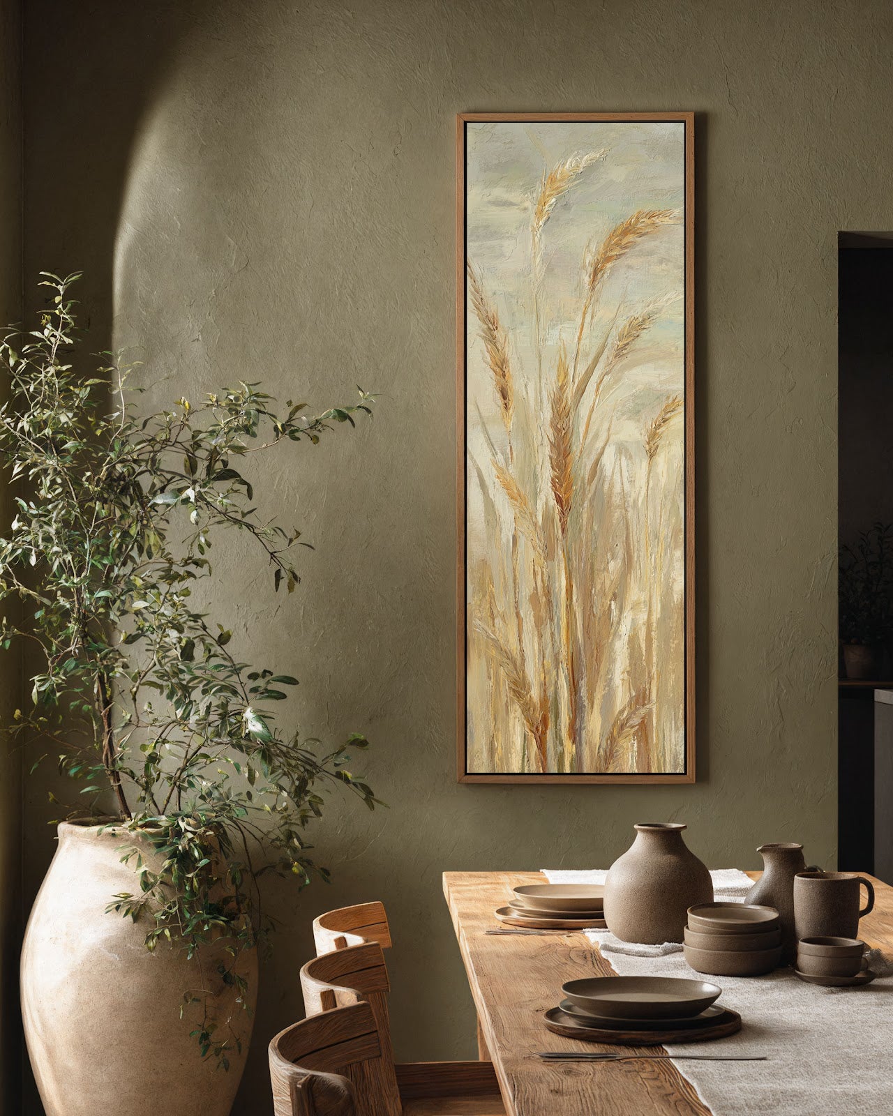 This vertical canvas print of impressionistic wheat stalks in warm earth tones is displayed in an oak floater frame on a textured olive wall above a wooden dining table.