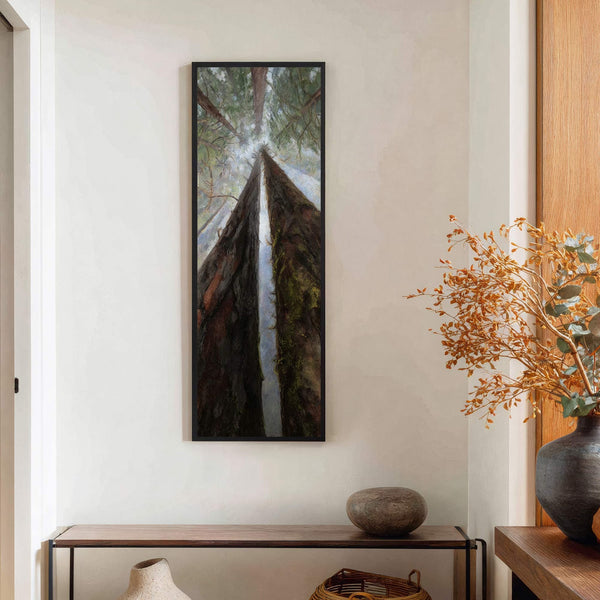 A vertical, oil painting print of redwood trees looking upwards, featuring earthy greens and browns, is framed in black and hangs on a white wall above a wooden console table.
