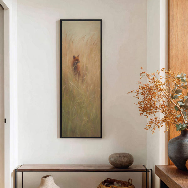 This vertical, impressionistic art print depicts a fox peeking through tall, golden grasses. The artwork is framed in black and hangs on a textured, cream-colored wall above a narrow wooden.