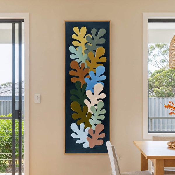 A vertical canvas wall art print featuring abstract leaf shapes in muted earth tones on a dark background, framed in natural wood and displayed on a beige wall next to.