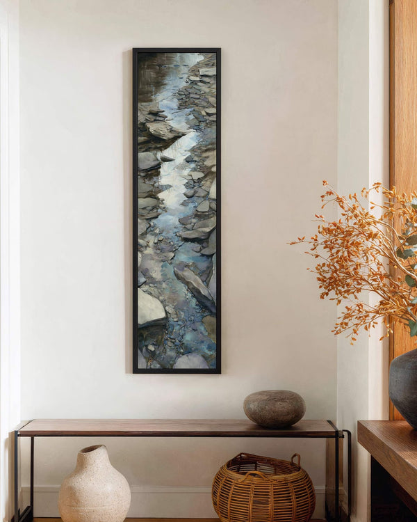 This vertical painterly canvas print depicts a blue and grey rocky stream in a black floater frame, displayed on a white hallway wall above a wooden console table.