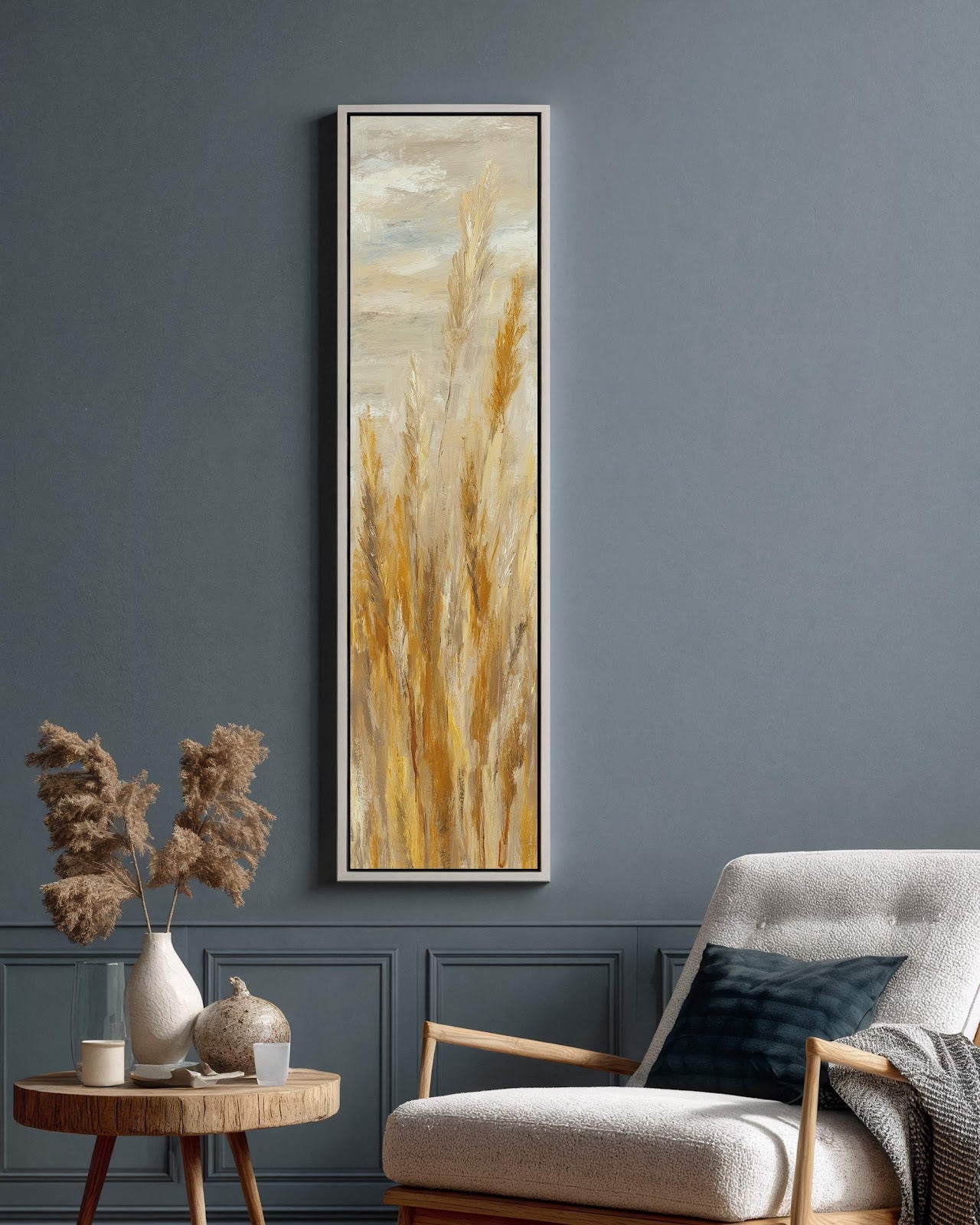 This vertical impressionistic canvas print depicts golden grasses in a white floater frame, displayed in a living room with dark blue paneled walls and a white armchair.