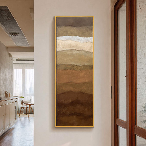 This vertical canvas print features abstract, wavy layers in a warm, earthy palette of browns and creams, framed with a thin gold border. It hangs on a light beige wall.