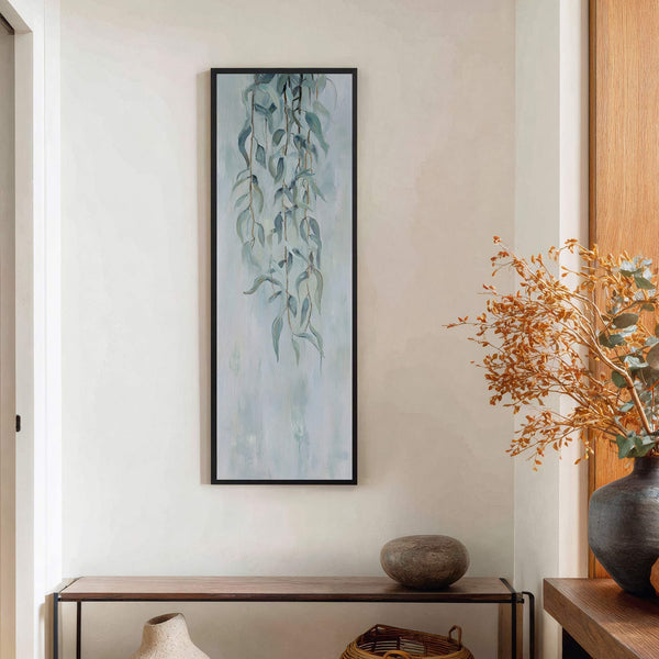 This vertical canvas print features a delicate painting of hanging eucalyptus leaves in a pale blue and green palette, framed in black and displayed above a wooden console table.
