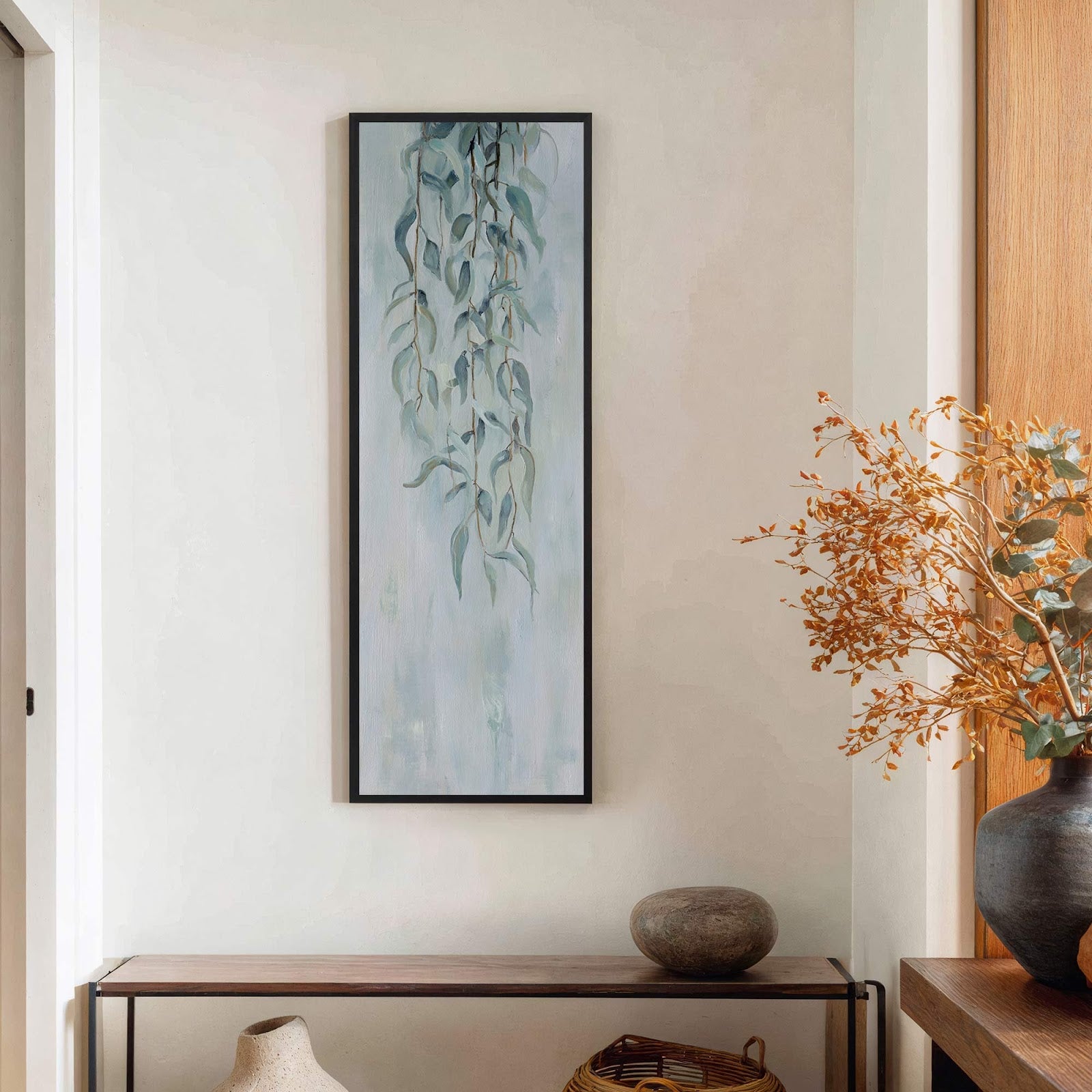 This vertical canvas print features a delicate painting of hanging eucalyptus leaves in a pale blue and green palette, framed in black and displayed above a wooden console table.