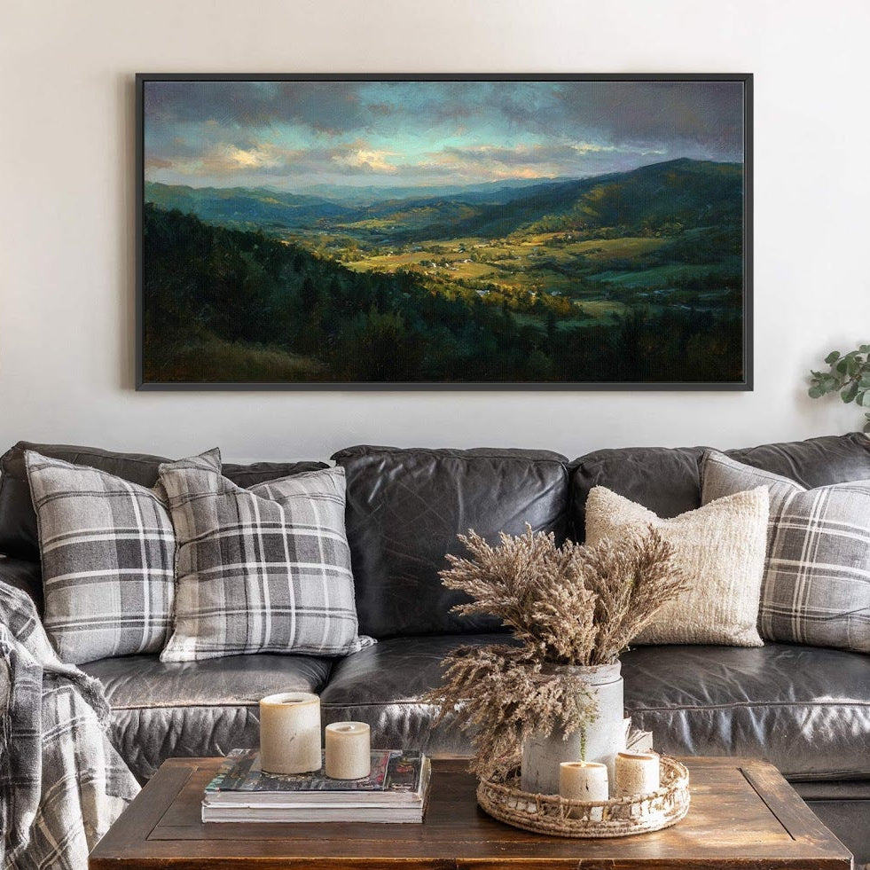 This horizontal canvas wall art displays a vibrant landscape painting with a palette of greens and blues, depicting rolling hills and a village, displayed above a dark leather sofa with.