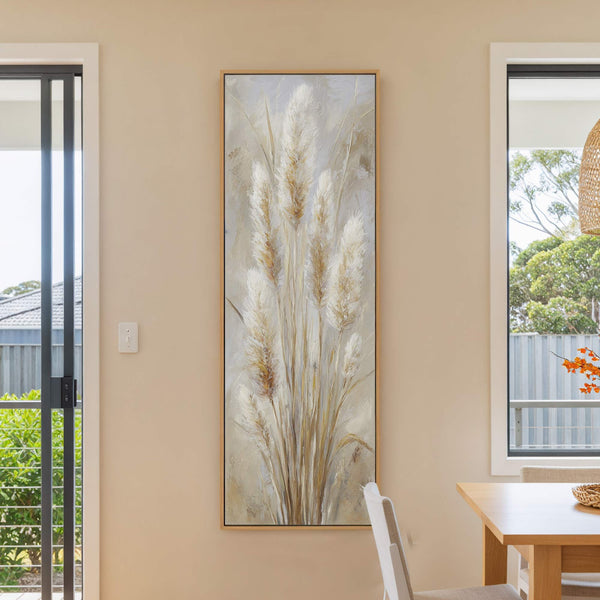 This vertical, impressionistic canvas wall art features tall, feathery pampas grass in a neutral palette, set against a light beige wall near a dining table.