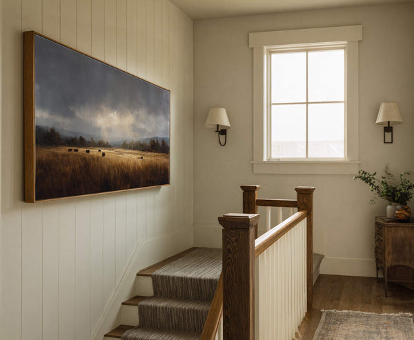 This horizontal canvas wall art features a moody landscape with dark skies over a golden field and hay bales, presented in a natural wood floater frame on a white paneled.