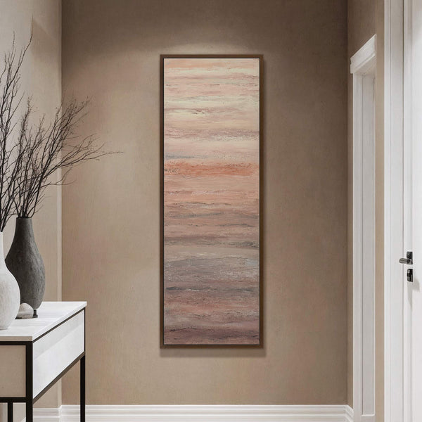 This vertical canvas wall art features an abstract, textured landscape with a palette of soft pinks, peaches, and grays, framed in dark walnut, displayed in a hallway with taupe walls.