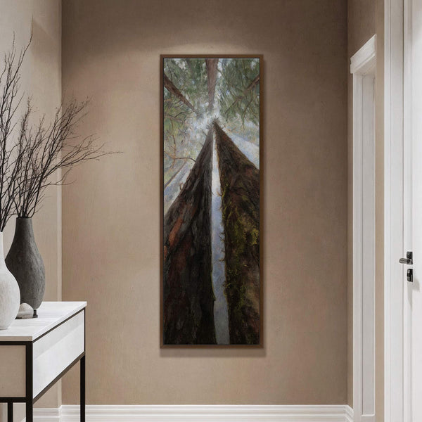 This vertical canvas wall art features a dramatic low-angle view of towering trees with dark bark and green moss, set against a pale sky, framed in walnut and displayed on.