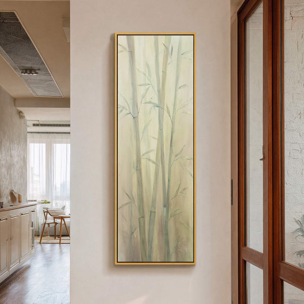 A vertical canvas print depicts a serene bamboo forest in a soft, muted palette with a golden frame, hanging on a pale wall in a modern room near a wooden.