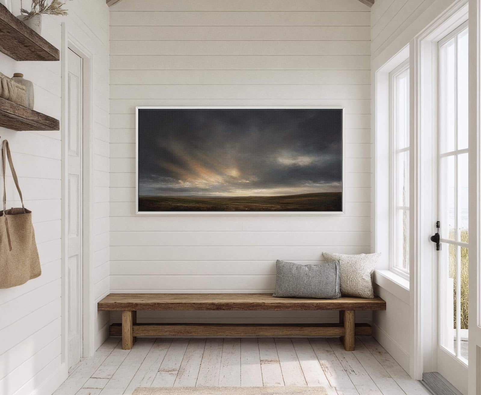 This horizontal, modern canvas wall art features a dramatic, moody landscape with dark clouds and a sunlit horizon, displayed in a white floater frame above a rustic wooden bench in.