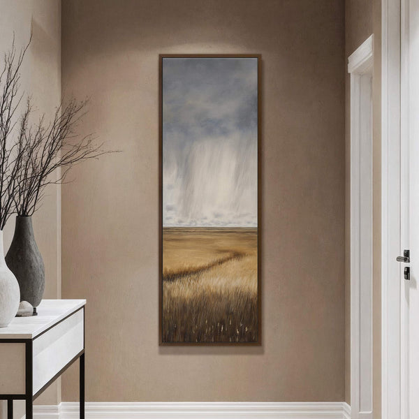 A vertical, impressionistic art print depicts a golden field under a stormy, pale blue sky, framed in dark wood and displayed in a hallway with a console table.