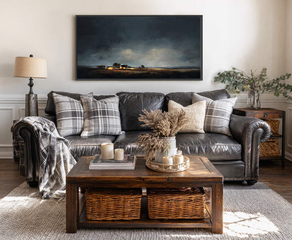 This framed canvas wall art features a dark, moody landscape painting with a soft glow from distant buildings, displayed above a black leather sofa with plaid pillows.
