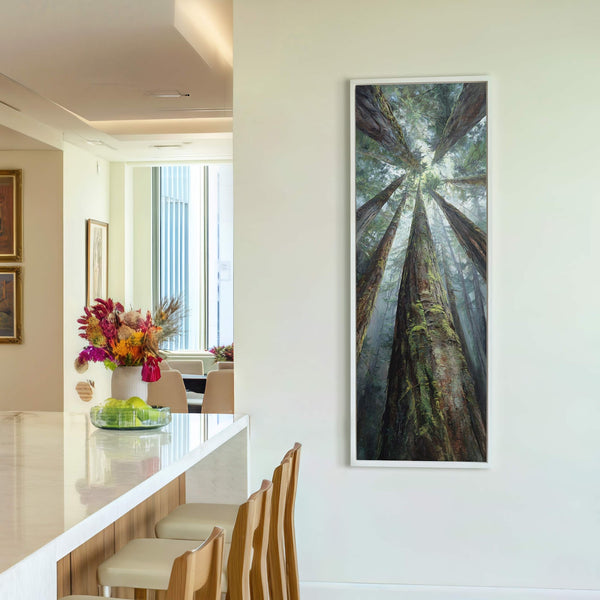 This vertical canvas wall art depicts a realistic forest scene with tall trees looking upwards. The artwork is framed in white and hangs on a plain white wall next to.