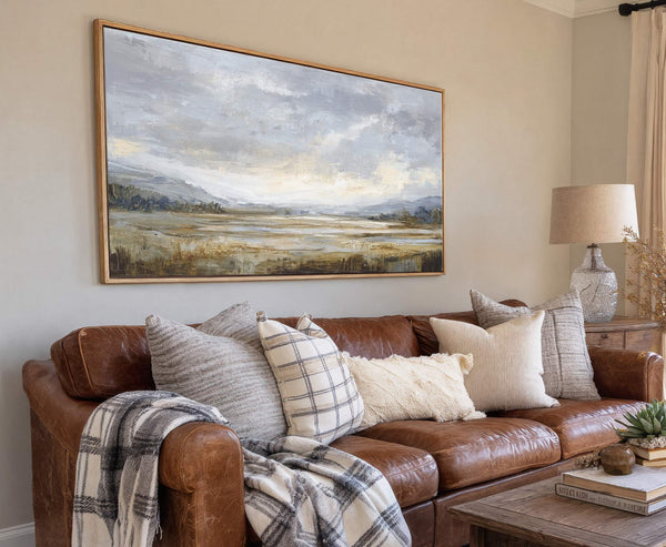 This horizontal canvas wall art features an abstract landscape painting in muted blues, grays, and earthy tones, displayed above a brown leather sofa with several decorative pillows in a living.