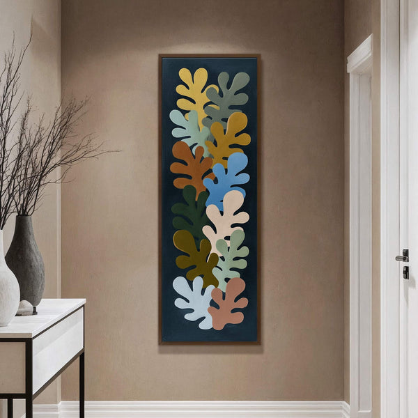 This vertical art print features a collection of abstract leaf shapes in muted yellow, blue, green, and brown tones against a dark background, displayed on a taupe wall next to.