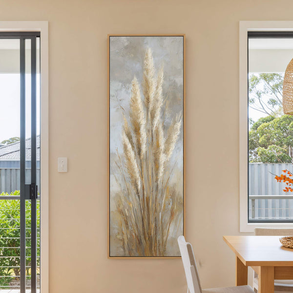 A vertical canvas wall art print features a textured, impressionistic painting of tall, golden pampas grass against a neutral, abstract background, framed in natural wood and displayed on a beige.