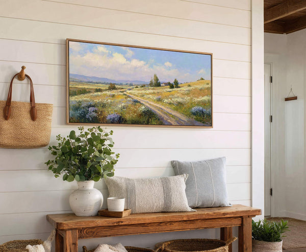 A horizontal canvas wall art print displays a landscape painting of a dirt road winding through a field of wildflowers, with distant mountains and a house. The artwork is framed.