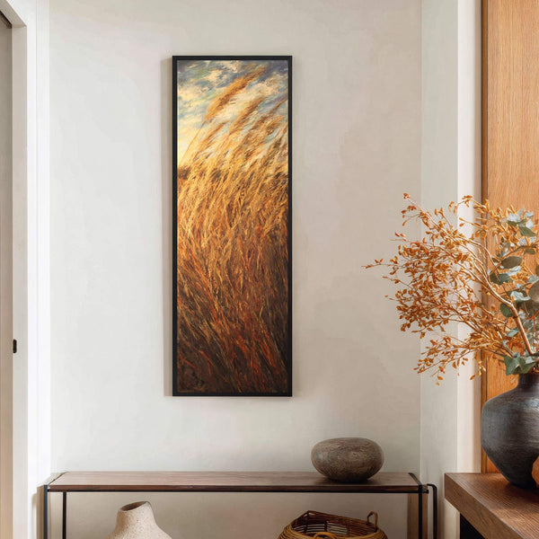 Vertical art print in a black frame depicts a textured, warm palette of golden reeds against a sky, displayed on a light-colored wall above a console table with a vase.