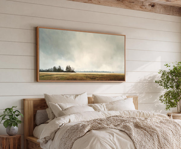 This horizontal landscape painting print features a muted palette depicting a field with trees under a cloudy sky, framed in natural wood and displayed above a bed on a white.