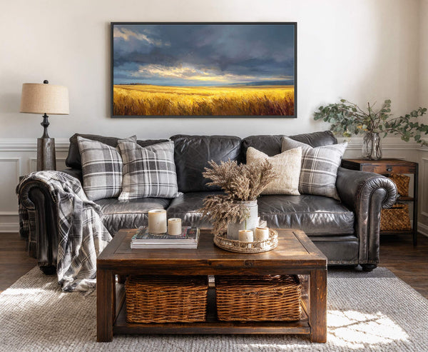 This horizontal canvas wall art depicts a golden wheat field under a dramatic, cloudy sky. The impressionistic style and warm palette are displayed on a neutral wall above a dark.